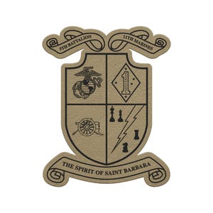 May include: A tan-colored emblem featuring the text "5th Battalion" and "11th Marines" on scrolled banners. The emblem includes a shield design with various military symbols and the text "THE SPIRIT OF SAINT BARBARA".