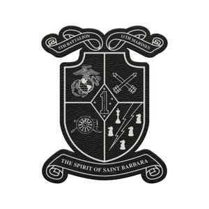 May include: A black and white emblem featuring the text "5th Battalion" and "11th Marines" at the top. The emblem has a shield shape with various symbols, including a globe, crossed cannons, and the text "THE SPIRIT OF SAINT BARBARA".