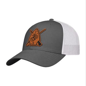 May include: A gray baseball cap with a white mesh back. The cap features a brown leather patch with a military emblem, including crossed rifles and the number "8". The brim is black.