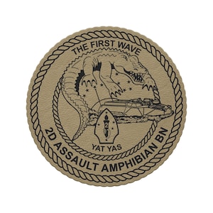 May include: A circular tan patch with a black rope border. The center features a black graphic of an alligator above an amphibious vehicle, with the text THE FIRST WAVE, 2D ASSAULT AMPHIBIAN BN, and YAT YAS surrounding a torch emblem.
