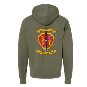 May include: A green hooded sweatshirt with a military-inspired patch on the back. The patch features a red shield with a knife and the text "3/7" in white. The text "2D BATTALION 7TH MARINES" is above the shield, and "THE CUTTING EDGE" is below the shield.