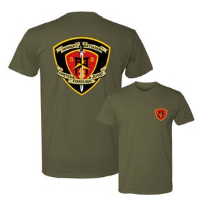 May include: Two olive green t-shirts. The larger shirt features a shield design with the text "AMERICA'S BATTALION", "FORTES", "JUVAT", and "FORTUNA". The smaller shirt has a smaller emblem on the chest.