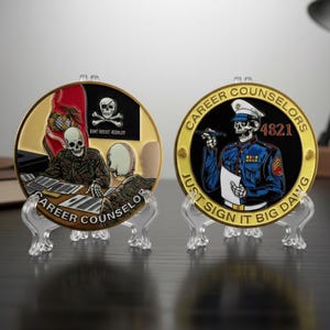 May include: Two gold-colored challenge coins on clear stands. One coin features a skull in a military uniform holding a gun and a paper, with the text "CAREER COUNSELORS" and "JUST SIGN IT BIG DAWG". The other coin has a skull and the text "CAREER COUNSELOR".