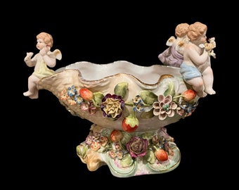 May include: A porcelain bowl with a shell-like shape, decorated with flowers and cherubs. The bowl is on a pedestal base and has a gold trim.