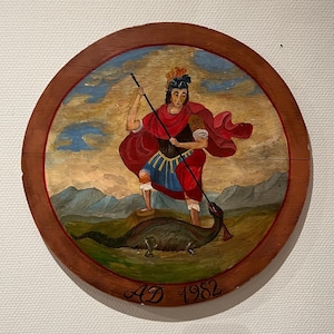 Puede incluir: Una placa redonda de madera con una imagen pintada de una persona vestida de rojo y azul de pie sobre un dragón verde. La persona sostiene una lanza y lleva una corona en la cabeza. La placa tiene el texto "AD 1982" escrito en ella.