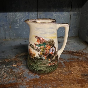 Może przedstawiać: Wazon ceramiczny w stylu vintage z uchwytem, z kolorową ilustracją postaci w krajobrazie. Wazon ma biały korpus, brązową krawędź i zieloną podstawę, ustawiony na rustykalnej drewnianej powierzchni.