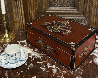May include: A wooden box with hand-painted floral designs in red, blue, yellow, and green. The box is closed and sitting on a marble surface. A white candle and a gold candlestick are to the left of the box. A white teacup and saucer with blue floral designs are to the right of the box.
