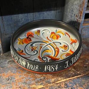 Könnte beinhalten: Ein schwarz-weiß bemaltes Holztablett mit einem floralen Design in Orange, Gelb und Blau. Das Tablett trägt den Text "God til ditt hus - 1987 - J. Toland" am Rand.