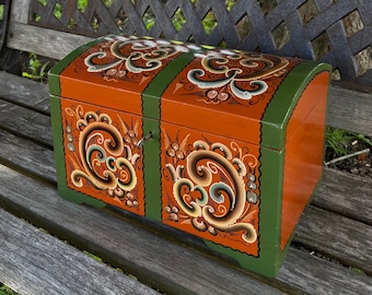 May include: A wooden box with a hinged lid, painted in a traditional folk art style with orange, green, and brown floral designs. The box is sitting on a wooden bench.