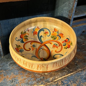 Könnte beinhalten: Eine große, runde Holzschale mit handbemaltem Blumendesign in Orange-, Gelb-, Blau- und Grüntönen. Die Schale hat einen braunen Rand und die Inschrift "DMMAR HINC 1981" in orangefarbener Schrift.