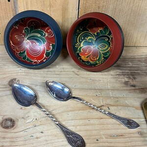 May include: Two wooden bowls with hand-painted floral designs in red, green, and yellow. The bowls are nested inside each other. Two silver spoons with twisted handles are on a wooden surface.