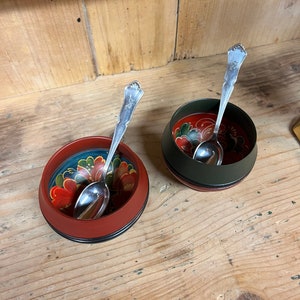 以下が含まれることがあります： 花柄が描かれた黒い縁取りの小さな木製のボウルが2つ。それぞれのボウルの中に銀のスプーンが入っています。