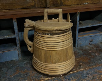Puede incluir: Un cubo de madera con tapa y asa. El cubo está hecho de madera natural y tiene un aspecto rústico. Está sentado sobre una superficie de madera.