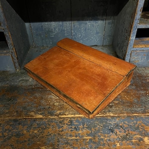 Op de afbeelding: Een houten bureau met een scharnierend deksel, geschilderd in een rijke oranje kleur. Het bureau staat op een blauw en bruin houten oppervlak.