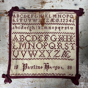 以下が含まれることがあります： 赤い刺繍のアルファベット、数字、そして「Pauline Nagel」という名前が1888年に日付が付けられた白いサンプラーです。