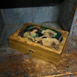 Peut inclure: Une boîte en bois vintage avec une finition dorée et un couvercle décoratif représentant une illustration de deux femmes portant des chapeaux. La boîte a un trou de serrure et semble vieillie.