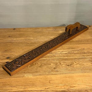 Pode incluir: Uma bandeja de madeira retangular e longa com uma inserção de couro esculpido. O couro apresenta um padrão floral detalhado. Uma pequena alça de madeira é fixada em uma extremidade. A bandeja é exibida em uma superfície de madeira.