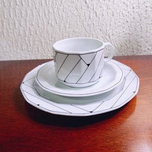 Puede incluir: Un juego de taza y platillo de cerámica blanca con un patrón geométrico en blanco y negro. El platillo tiene un borde festoneado.