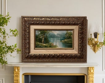 May include: A framed landscape painting depicting a river scene with trees and a waterfall. The painting is in a brown ornate frame with a gold trim.