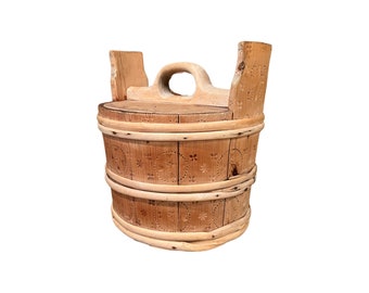 Puede incluir: Un cubo de madera con tapa y asa. El cubo está hecho de madera marrón claro y tiene un diseño tallado en el lateral. El cubo está sobre un fondo blanco.