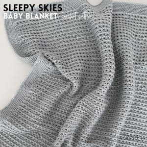 May include: A gray, crocheted baby blanket with the text "SLEEPY SKIES BABY BLANKET crochet pattern" in the upper left corner. The blanket has a textured, open weave design and is folded, showing the details of the stitch work.
