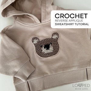 May include: A beige hooded sweatshirt with a crocheted bear applique. The bear is brown with cream-colored accents. The text "CROCHET REVERSE APPLIQUE SWEATSHIRT TUTORIAL" is displayed in a white rectangle. The sweatshirt has ribbed cuffs and hem.