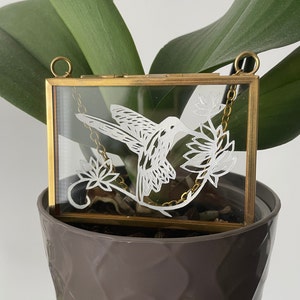 May include: A rectangular glass frame with a thin brass metal border and two top rings for hanging. The glass features a white etched design of a hummingbird hovering near two stylized flowers, with a gold-toned chain visible inside the frame.