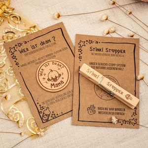 Könnte beinhalten: Zwei Holzprodukte für Babys. Das erste ist ein runder Holztoken mit dem Text "Wer ist dran?" und "Mama" mit einem Baby-Cartoon darauf. Das zweite ist eine Holzklammer mit dem Text "Stinki Stopper" und "Noch nie war Windeln wechseln so einfach".