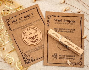Könnte beinhalten: Zwei Holzprodukte für Babys. Das erste ist ein runder Holztoken mit dem Text "Wer ist dran?" und "Mama" mit einem Baby-Cartoon darauf. Das zweite ist eine Holzklammer mit dem Text "Stinki Stopper" und "Noch nie war Windeln wechseln so einfach".