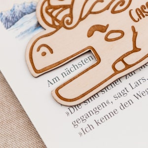 May include: A wooden bookmark shaped like a dragon with the text "Am n&auml;chsten" (German for "Next") visible. The bookmark is resting on a page of a book.