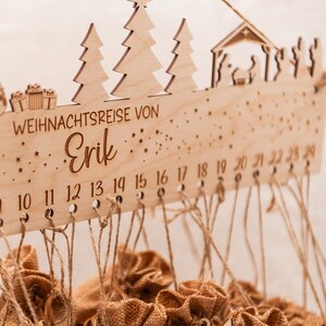 May include: Wooden Christmas advent calendar with a star-filled sky design. It features a nativity scene and two Christmas trees. The calendar has 24 numbered slots with twine and burlap bags hanging from each. The text "WEIHNACHTSREISE VON Erik" is engraved on the calendar.