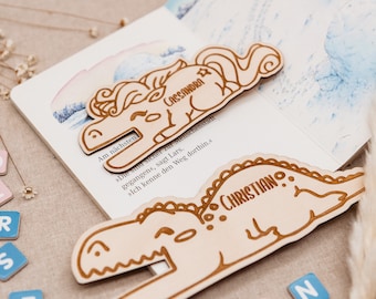 May include: Two wooden bookmarks, one shaped like a unicorn and the other like a dinosaur. The unicorn bookmark is light brown and has the name "CASSANDRA" engraved on it. The dinosaur bookmark is also light brown and has the name "CHRISTIAN" engraved on it.