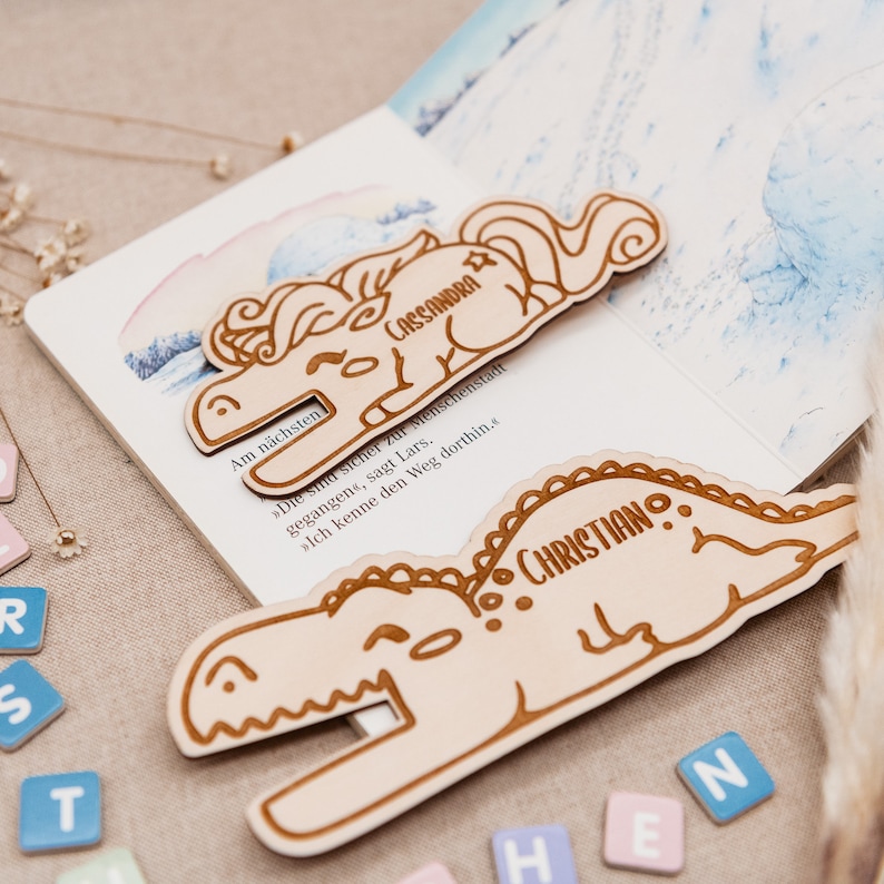May include: Two wooden bookmarks, one shaped like a unicorn and the other like a dinosaur. The unicorn bookmark is light brown and has the name "CASSANDRA" engraved on it. The dinosaur bookmark is also light brown and has the name "CHRISTIAN" engraved on it.
