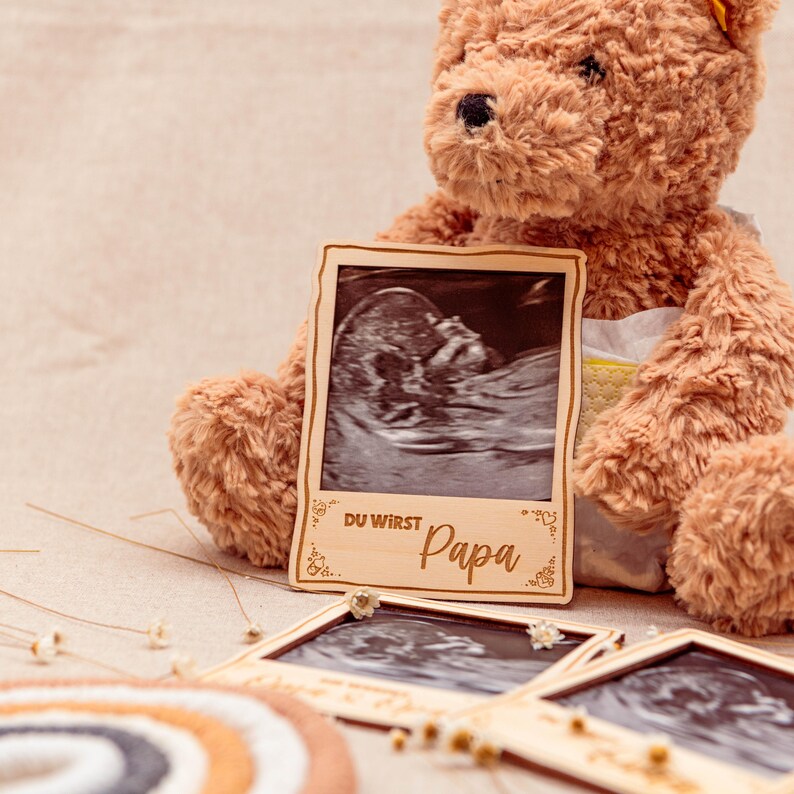 May include: A wooden photo frame with a black and white ultrasound image inside. The frame has a decorative border and the text "Du wirst Papa" in German.