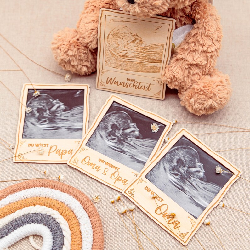 May include: Four wooden photo frames with ultrasound images and text. The frames are decorated with small white flowers and gold string. The text on the frames reads "Du wirst Papa", "Ihr werdet Oma & Opa", "Du wirst Oma", and "Dein Wunschtext".