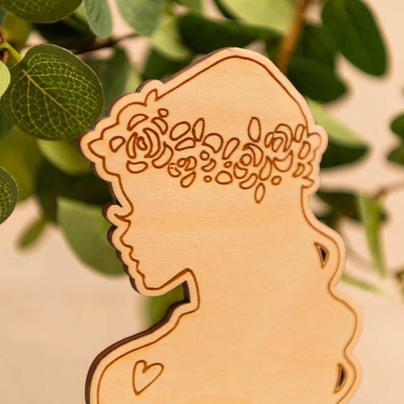 May include: Wooden silhouette of a woman's profile, decorated with a floral crown. The design is laser-cut from light-coloured wood, with a heart detail. The silhouette is set against a backdrop of green foliage.