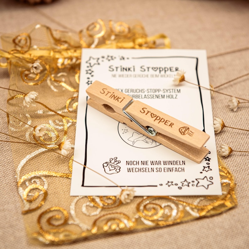 May include: A wooden clothespin with the words "Stinki Stopper" printed on it. The clothespin is on a white card with the text "Noch nie war Windeln wechseln so einfach" printed on it.