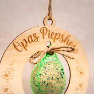 Pode incluir: Um comedouro de p&aacute;ssaros de madeira com o texto "Opas Piepshow" gravado nele. Um saco de malha verde cheio de sementes para p&aacute;ssaros pendura no comedouro.