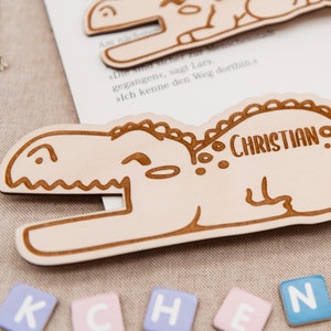 May include: A light brown wooden dinosaur bookmark with the name "CHRISTIAN" engraved on it. The bookmark is lying on a beige fabric surface with colourful letter blocks spelling out "KCHEN" in front of it.