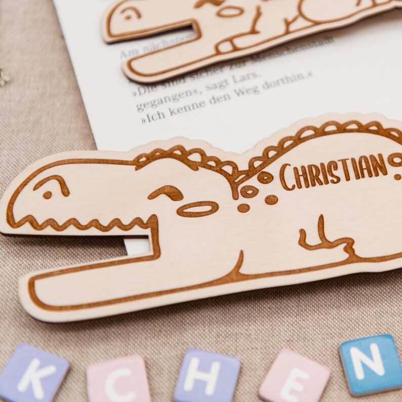 May include: A light brown wooden dinosaur bookmark with the name "CHRISTIAN" engraved on it. The bookmark is lying on a beige fabric surface with colourful letter blocks spelling out "KCHEN" in front of it.