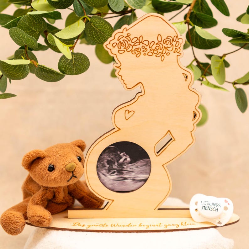 May include: A wooden pregnancy announcement frame shaped like a pregnant woman, displaying a sonogram image. A brown teddy bear and a pacifier with text are next to the frame. The frame has the text "Das grö&szlig;te Wunder beginnt ganz klein."