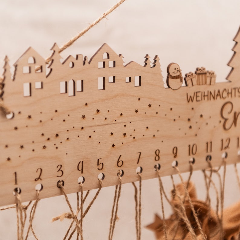 May include: Wooden Christmas advent calendar with a town silhouette, stars, and numbers 1 through 12. The calendar has a string attached to each number, which can be used to hang ornaments or other decorations.