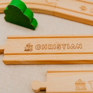 May include: Wooden train track piece engraved with the name "CHRISTIAN". A green wooden tree is in the background. The track is light brown and features a train graphic. Other track pieces are visible.