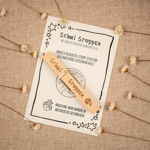 May include: A wooden clothespin with the words "Stinki Stopper" printed on it. The clothespin is on a white card with the text "Nie wieder Ger&uuml;che beim Wickeln" and "Unisex Geruchs-Stopp-System aus Naturbelassenem Holz".