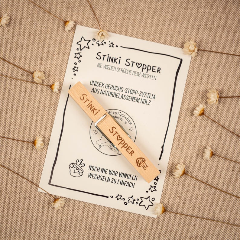 May include: A wooden clothespin with the words "Stinki Stopper" printed on it. The clothespin is on a white card with the text "Nie wieder Ger&uuml;che beim Wickeln" and "Unisex Geruchs-Stopp-System aus Naturbelassenem Holz".