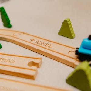 May include: A wooden toy train track with the name "KARL" engraved on a curved section. The track is surrounded by green wooden tree-shaped figures and a blue toy train. The image evokes a sense of childhood and play.