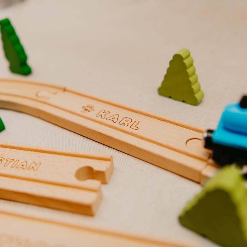 May include: A wooden toy train track with the name "KARL" engraved on a curved section. The track is surrounded by green wooden tree-shaped figures and a blue toy train. The image evokes a sense of childhood and play.