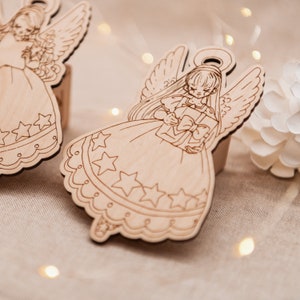 Peut inclure: Deux d&eacute;corations d'ange en bois avec des ailes, des robes et des &eacute;toiles. Un ange tient une bo&icirc;te cadeau avec un n&oelig;ud.