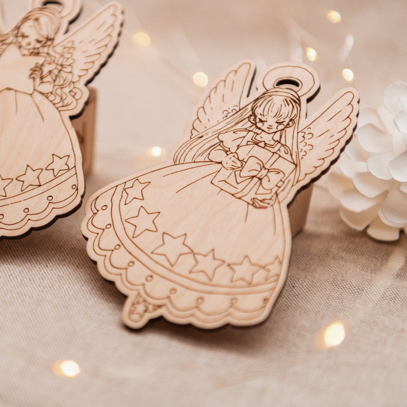 Peut inclure: Deux d&eacute;corations d'ange en bois avec des ailes, des robes et des &eacute;toiles. Un ange tient une bo&icirc;te cadeau avec un n&oelig;ud.