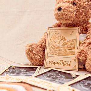 May include: A wooden photo frame with a brown teddy bear holding it. The frame has a cutout of an ultrasound image and the text "Dein Wunschtext".